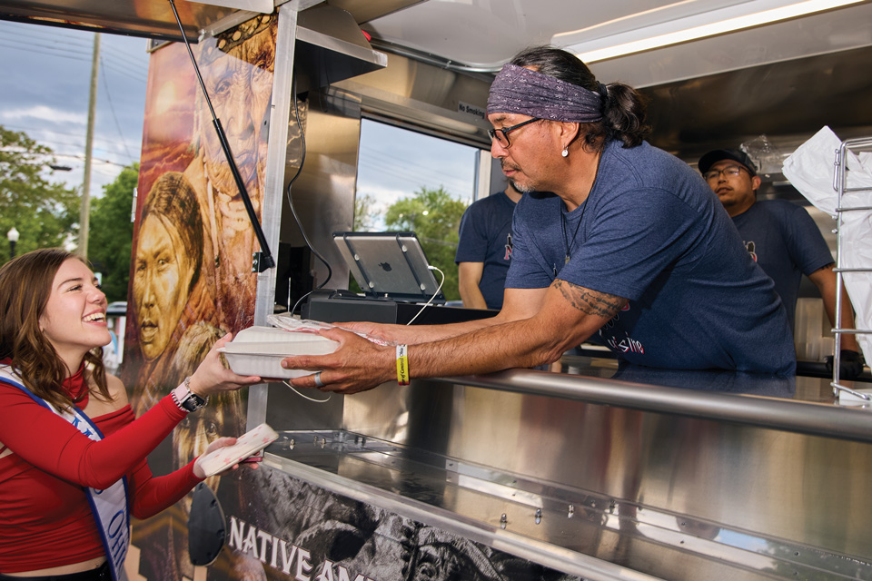 Ty Smith of NAICCO Cuisine’s food truck handing customer box of food (photo by Brian Kaiser)