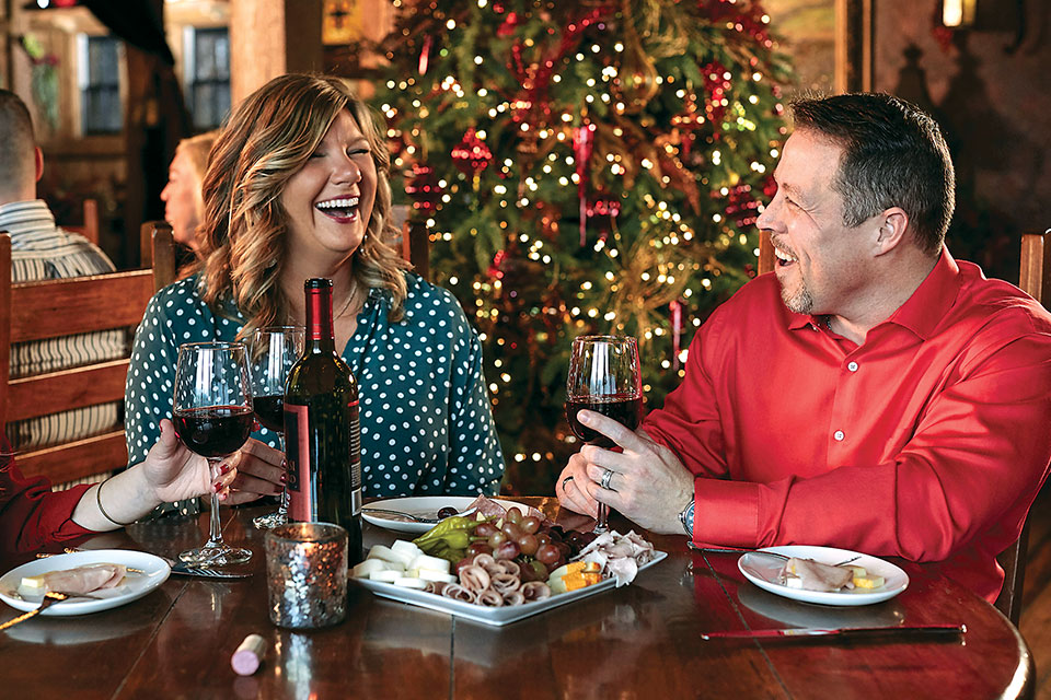 A couple laugh over a dinner with wine and charcuterie in front of a Christmas tree.
