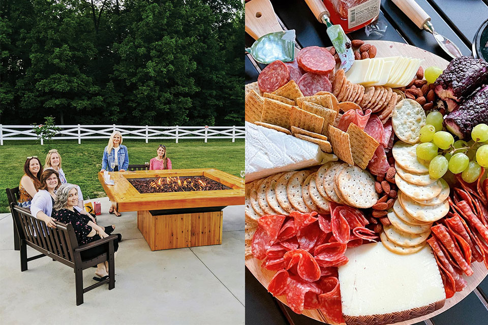 A group of friends relax by a fire pit outside on a summer night. Next to it, a charcuterie spread.