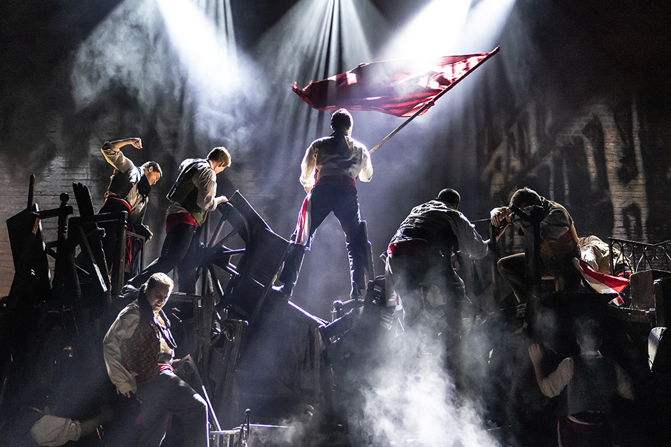 Performers standing on barricade set while waving flag in Les Miserables (photo by Matthew Murphy)